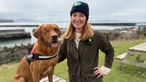 A fox red Labrador is sitting on top of a picnic bench. One of his eyes has been sewn shut. His mouth is open and his tongue is sticking out. A woman has one hand on his back and the other hand on her hip. She's wearing a hat that says RSPB.