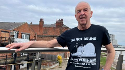 Paul Roberts has cropped silver hair and is standing on a bridge showing off a black t shirt that has a dazed, fallen down elephant on it along with the words "I'm not drunk, I have Parkinson's" written on. In smaller letters beneath it says "okay...maybe I'm a 'lil drunk. #parkinson's-awareness"