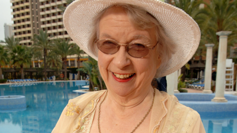 Elsie King has blonde/grey hair, white sun hat, yellow dress, sunglasses and a chain around her neck.