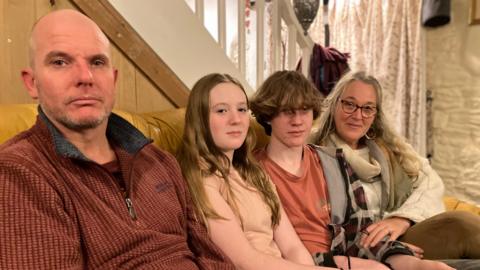 The family sit on a sofa in the living room of their small rented cottage. Rob is closest to the camera, wearing a dark red fleece, while next to him to his right sits Olive, his daughter, then Fin, his son, with their mother Joanna sitting at the end. All look toward the camera looking serious, except Jo who smiles slightly.