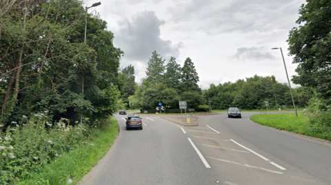 Cars driving towards and away from a roundabout with greenery on either side of the road