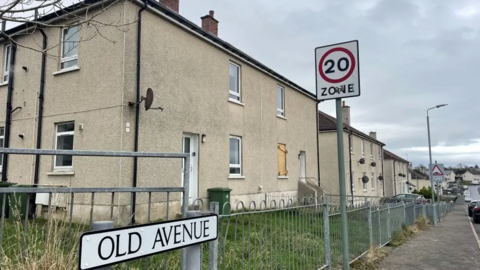 Old Avenue in Auchinleck. A sign with the name of the street is in the foreground. Behind it is a grey metal fence. Behind that is a light-coloured block of flats. One of the windows is boarded up with light-coloured wooden chipboard.