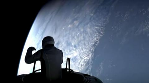 A silhouette of someone in a spacesuit who is stretched out of their capsule from the waist up in space, overlooking the Earth