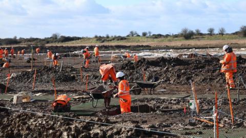 Lots of workers in orange hi-vis outfits work on a very muddy site at Coalhouse Fort. They are using spades, wheelbarrows and rope.