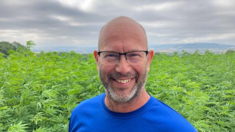 Jake Freestone pictured standing in front of a field of hemp. He has a bald head and grey stubble. He is wearing think glasses and a blue t-shirt.