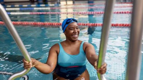 A woman wearing a blue swim cap and goggles holds the handrails while stepping into an indoor swimming pool, with lane markings visible in the water behind them.
