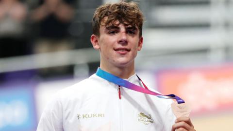 Henry Hobbs, wearing Team England's white and red tracksuit jacket, holds up his men's elimination race bronze medal
