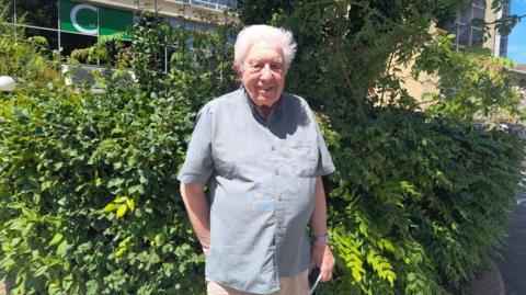 Keith Taylor, 84, with short white hair, wearing a pale blue shirt and beige chino trousers. He is standing in front of a bush on a sunny day and smiling at the camera.