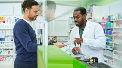 A stock photo of a male customer talking to a male pharmacist at a counter in a shop. Behind them both, on each side of the counter which has a screen, are shelves of medicines. The customer's face is reflected in the screen. 