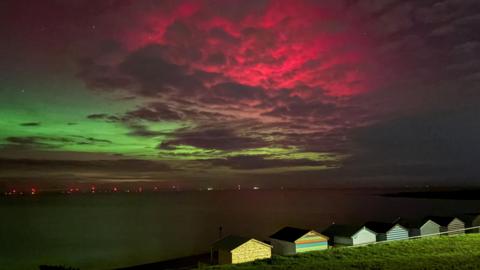 An amateur photograph of the Northern Lights. There are pink and green lights in the sky above the sea.