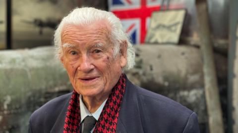 Gordon Talbot sits in the museum talking to the BBC. He is wearing a shirt and tie with a navy jacket and a red scarf. There is a union flag behind him.