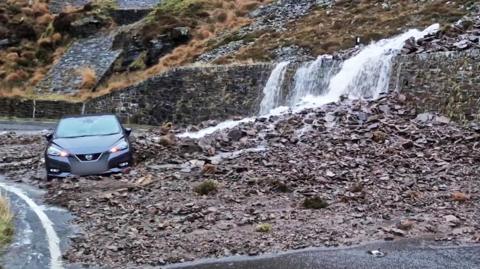 A car sits on the side of the road as debris from a landslip hits