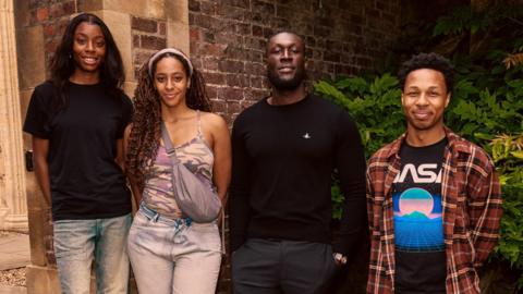 The rapper Stormzy with student Destiny, Drew and Dimario alongside him. They stand outside a brick building with shrubbery.