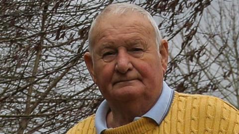 Trevor Smith, who has white hair, is wearing a yellow jumper. Trees are behind him.