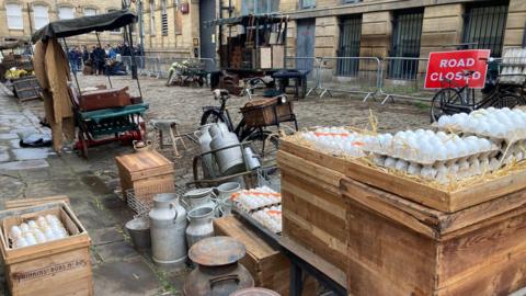 White eggs stacked in row on top of wooden boxes and metal milk churns on a cobbled street.  