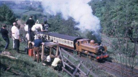 A mustard brown-coloured locomotive pulling two carriages with a film crew looking on from a hillside