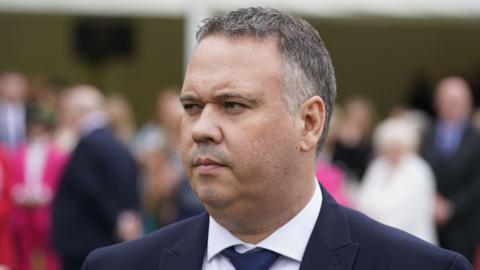 A close up image of John Caldwell, wearing a white shirt and navy suit. He has short grey hair. The background of the image is blurry. 