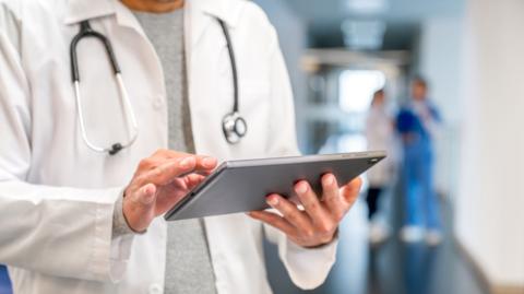 A doctor in a white medical coat reads from a tablet computer.