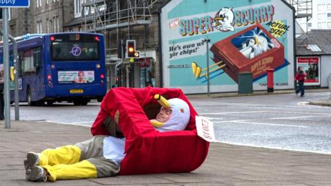 Man in a seagull costume, lying on the pavement, with an artwork of a seagull in the background on a wall that he is recreating.