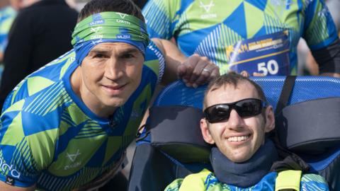 Kevin Sinfield (left) and Rob Burrow (right) wearing rugby kit. Sinfield is leaning towards Burrow's adjustable chair. Both men are smiling at the camera.