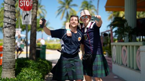 Scotland fans in kilts dancing in the street, with palm trees and the Miami beach in the background