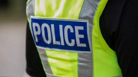The back of a British police vest. It is illuminous yellow with silver reflective strips on the side. It says POLICE is bold white lettering on a blue rectangular background 