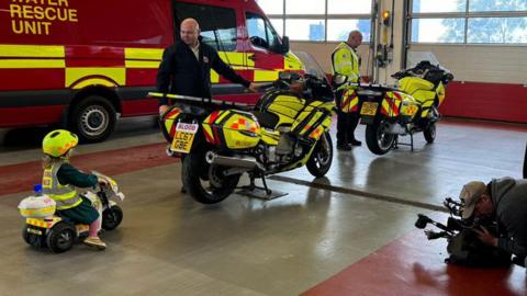 Three-year-old Beatrix dressed in rescue uniform and riding a toy emergency service bike behind two blood bikers, standing next to two motorbikes and also dressed in florescent emergency clothing.