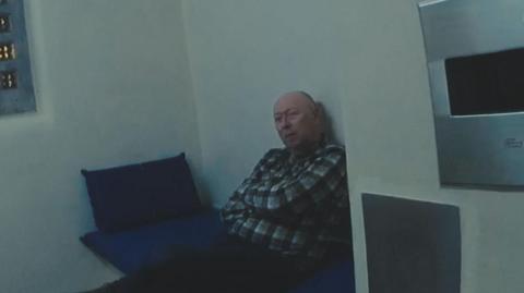 Stephen Alderton is wearing black trousers and a checked shirt, and is sitting in a police cell on a bed with a blue pillow and mat. There is the edge of a window on the left.