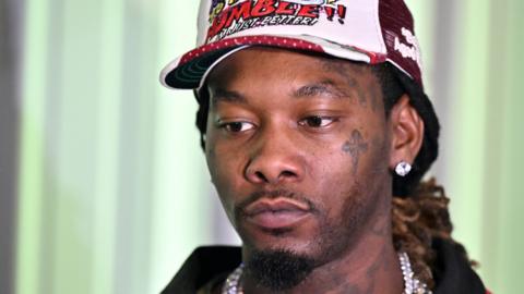 Offset is a man, he has brown eyes and is wearing a white and red cap. He has a diamond stud earring and is wearing a large silver chain. He is wearing a red jacket with a black collar. He has a tattoo on his cheek below his eye of a cross and tattoos on his neck.