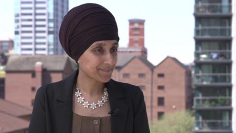 Sukhvinder Kaur is wearing a dark Sikh head covering or turban. He has a black jacket, with brown top and a striking necklace of white flowers. 