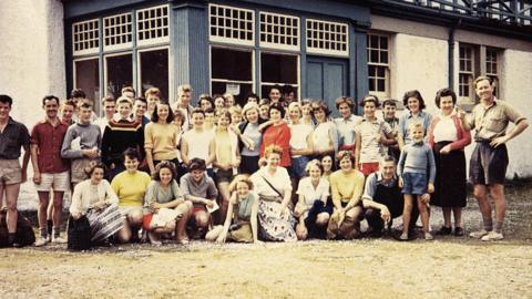 A colour picture of a large group of people standing in sunshine outside a large white-walled building with a blue-painted porch. The group is made of men, women and children. Some of them are wearing shorts and short sleeves.