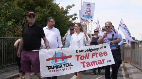 A group of people crossing the bridge. At the front are three men and one woman. They are holding picket signs and a banner which says Campaigning for a toll-free tamar. 