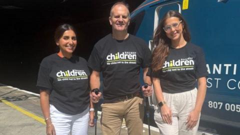 Two women and a man wearing black t-shirts with a white and green Railway Children writing and log on the front. They are standing on a station platform in front of a blue train. The woman on the left has black hair and white trousers and is smiling. The taller man has hiking poles in each hand and is wearing beige trousers. The woman on the right is younger, has long dark hair and transparent framed glasses. She is wearing beige trousers.