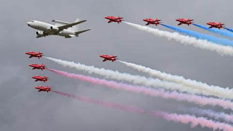The Red Arrows flying in formation, with red and white smoke trailing behind. Also flying alongside them is a larger white jet-style plane.