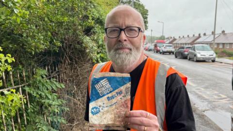 A man in high vis holding up a crisp packet with blue and white design on it and a 15p price