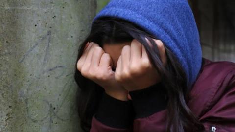 A teenage girl obscures her face with balled fists pressed against her eyes as she leans against a wall.