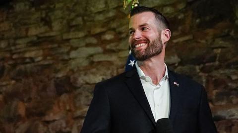 Clay Fuller, in a suit jacket and white collared shirt, looks to the side and smiles
