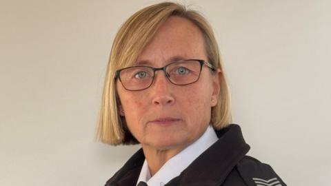 Laura Heggie, who has short blonde hair and wears black rimmed glasses. She is wearing her police uniform, consisting of a white shirt and a black coat that has epaulettes on.