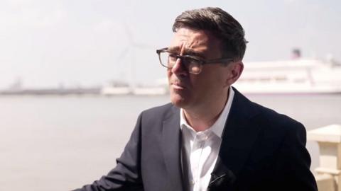 Andy Burnham pictured in front of a coastline wearing a white shirt and black suit jacket