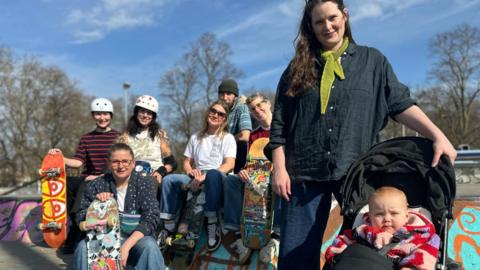 A group of mums sit together at a skate park. One mum has a baby in a pram. 