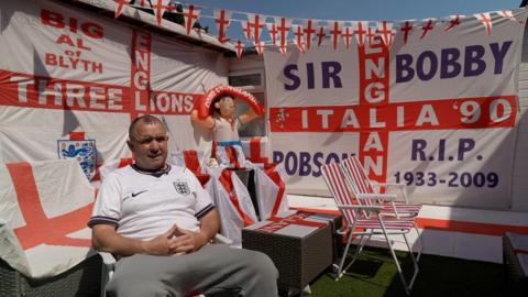 Alfie Patterson, a middle-aged man wearing an England football shirt, sitting in his garden. England flags are draped on the walls and across the chairs. They are also are draped over the tables and from bunting above him. An England flag has been painted on the decking.