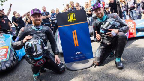 Ryan and Callum Crowe Kneeling next to a blue sign with the number one on it in orange. They are both wearing black leathers with blue trim and purple caps. They are holding motorcycle helmets that match their leathers.
