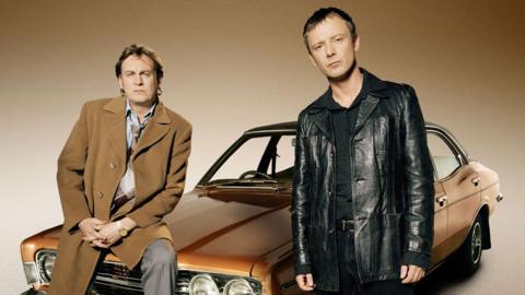 Promo shot of Life on Mars. Two men standing in front of brown car.