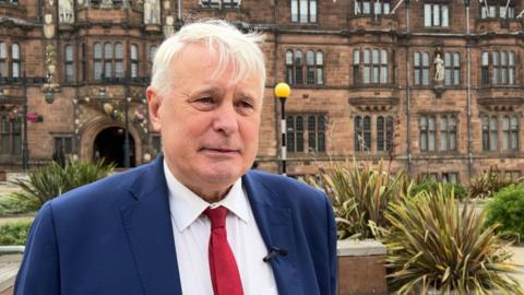 A man with white hair blown slightly over his forehead stands in front of a stone brick building with dozens of arched windows and plants between him and the building. He has a blue suit, white shirt and red tie on and a microphone clipped to a lapel.