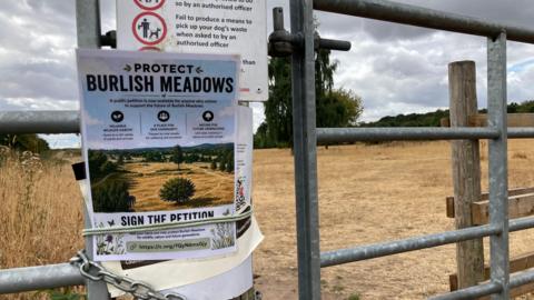 A poster that reads "protect Burlish Meadows" is pinned to a metal gate in front of a large open field that has turned brown from the dry weather.