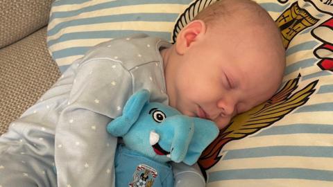 A newborn baby wearing a blue onesie with white stars lies on a blue and white stripy pillow, holding an elephant toy reading "Coventry City"