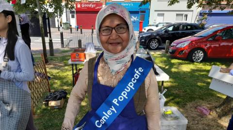 A woman standing in a park on a sunny day, shaded by the threes. She is wearing glasses and a headscarf. Over her clothes she has a blue tabard and a blue sash reading "Canley Popupcafe".