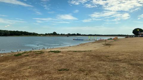 Alton Water is bordered by dried vegetation and dusty ground. There are green trees on the other side of the lake.