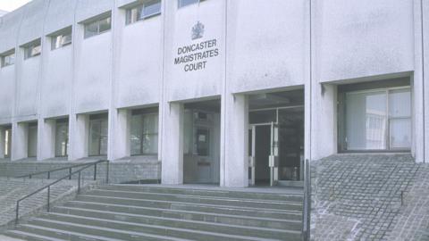 The exterior of a large, concrete building identified by the sign above the entrance as “Doncaster Magistrates’ Court”. The structure has a distinctly utilitarian, modernist design, with tall, rectangular pillars and evenly spaced windows along the upper level. A wide set of steps leads up to the main entrance, which has glass doors, one of which is standing open. To the right of the steps, the ground slopes upward in a textured ramp made of small, square paving stones. Metal railings run alongside the steps.
