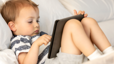 A toddler lying on his back in front of a screen, wearing grey shorts and a blue and white stripey tshirt. 